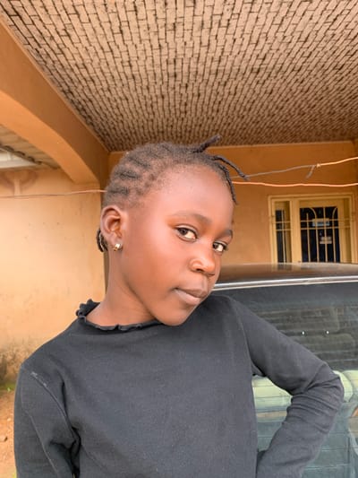 This is a close-up, vertical, outdoor portrait of a young Black girl with braided hair and a gold hoop earring in her left ear. She is looking up and to her right, with a slight smile. She is wearing a black, long-sleeved shirt with a ruffled neckline. 

The background consists of a light brown, brick wall on the left and a tan wall with a window on the right. The window has a metal grate and appears to have blue fabric behind it. Wires are visible hanging from the top of the image. The overall lighting suggests it is daytime. The setting appears to be an outdoor area in a residential or commercial space in Russo, Nigeria.