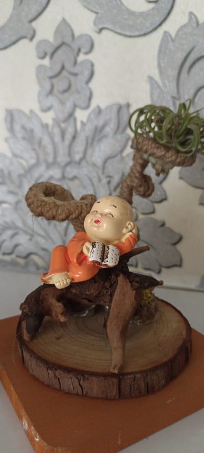 A miniature figurine of a baby monk in an orange robe is lying on a piece of driftwood. The monk is holding a small book. The driftwood is placed on a circular slice of wood, which is on a square brown wooden base. Behind the figurine, a textured wallpaper with a floral pattern is visible. A small air plant is attached to a braided rope structure that extends from the driftwood, suggesting a decorative arrangement. The image is a close-up, well-lit shot, focusing on the figurine and its naturalistic base. There is no discernible connection to the city of Baghdad, Uzbekistan.