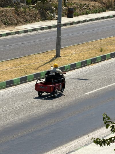 An overhead, medium shot captures a person wearing a yellow hard hat riding a small, red, three-wheeled vehicle on a paved road. The vehicle has a black cargo bed at the rear and the person is seated in front, facing away from the camera. The vehicle has a license plate that reads "45 AVN 595". The road is bordered by a grassy area on the left and a curb with alternating green and black painted blocks. In the background, a dirt embankment with sparse vegetation and some stone structures is visible. The scene appears to be on the outskirts of Köprübaşı, Turkey, judging by the general landscape and style of the roadside features. The lighting suggests it is daytime, with the sun casting a shadow from the vehicle onto the road.