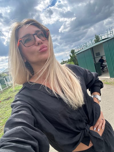 A young woman with long blonde hair and glasses is taking a selfie outdoors. She is wearing a black shirt tied at the waist and a watch on her left wrist. Her nails are painted a light blue color, and she is wearing a ring on her right hand. The woman has pink lipstick on and is making a pouty face. In the background, there is a green building with a metal roof and a fence. The sky is cloudy, suggesting it might be a partly cloudy day. The ground appears to be paved with asphalt or concrete and there is some grass visible. Another person is seen in the background, sitting on a bench inside the green structure. The image appears to have been taken during daylight hours, given the natural lighting. The overall impression is a casual outdoor selfie.