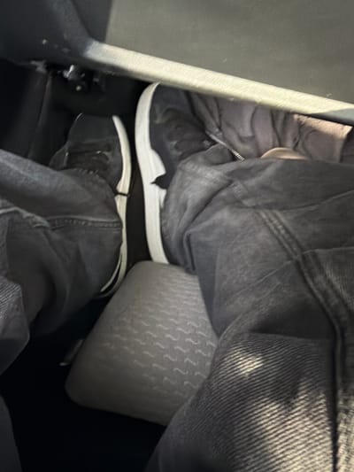 The image shows a close-up view of a person's legs and feet inside a vehicle, likely a car. The person is wearing dark denim jeans and black sneakers with white soles. The left leg is bent at the knee and the foot is resting on the car's floor, near what appears to be a pedal. The right leg is also bent and the foot is positioned slightly higher, with the sneaker visible.
The interior of the car is dark, with the dashboard and part of the seat visible in the background. There is a textured grey cushion on the floor in front of the passenger seat. The lighting suggests it is daytime, with some natural light coming into the car. The image is taken from a high angle, looking down at the person's legs. There is no indication of the specific location in Skumparp, Sweden.