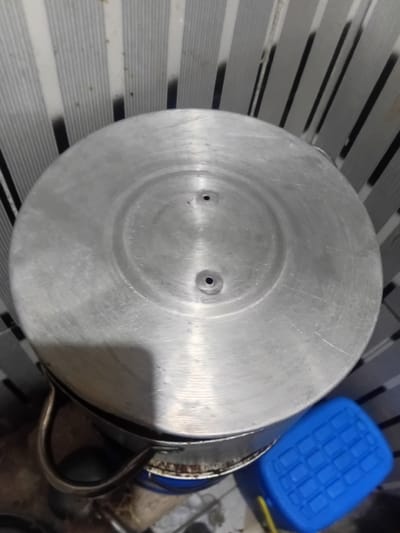 The image shows the top of a metal pot with two small holes in its lid. The pot is made of a brushed aluminum material and appears to be well-used, with visible scratches and scuffs. The lid has a concentric circular design pressed into it, and the two holes are positioned symmetrically within the inner circle. The pot has a metal handle attached to its side, which is also visible in the lower right portion of the image.

In the background, to the right of the pot, is a bright blue plastic container with a textured lid, reminiscent of a cooler or storage bin. A yellow object, possibly a piece of cloth or plastic, is visible near the blue container. The background also shows a striped surface, likely a floor or table, with alternating dark and light gray or white stripes. The overall scene appears to be indoors, given the lighting and the presence of household objects.

The image is taken from a high-angle, looking down at the pot and its surroundings. There is no indication of people, specific time of day, weather, or particular emotions. The location context of Pantinapu, Nigeria, does not provide any specific visual cues within the image itself, as the objects and setting are generic. No visible text is present in the image.