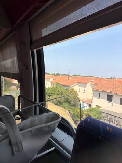 The image, taken from inside a vehicle, offers a view through a window of a residential area. The foreground is dominated by a partially visible white bag with handles and a metal rail. Beyond the immediate interior, the scene opens up to a landscape featuring multiple houses with distinctive orange tiled roofs. Lush green trees are interspersed between the buildings. The sky is clear and blue, suggesting a sunny day. A portion of a seat with a blue fabric cover is visible in the lower right of the frame, with graffiti reading "flair" on a concrete structure in the background. The context of Elne, France is provided, indicating the geographical location of this scene.