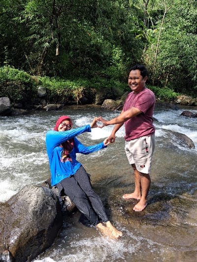 A man and a woman are standing in a shallow river, holding hands to form a heart shape. The man is wearing a maroon shirt and khaki shorts, while the woman wears a blue long-sleeved shirt, a red hijab, and dark pants. They both have bare feet. The river flows around rocks, and the river bank is lined with lush green vegetation. They are both smiling.