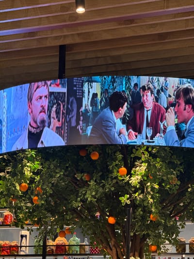 This media file captures an indoor setting, divided visually by a large vertical screen on the left and a decorative element on the right. The left portion of the image displays a tall, narrow screen showcasing scenes from what appears to be a film or television program. Within these displayed scenes, various people are visible, engaged in a social gathering. One man with dark, curly hair and a red cardigan smiles directly at the viewer, seated at a table with a wine glass. Another frame features a bearded man with a calm expression. The overall style of the on-screen content suggests a formal or period outdoor event.
Adjacent to the screen, on the right, stands an artificial tree adorned with abundant green leaves and numerous bright orange fruits, reminiscent of oranges or tangerines. The tree is artificially lit, casting highlights on its foliage and fruit. Behind the tree, shelves are visible, stocked with a variety of items such as clear glass bottles (some with green liquid), jars containing red contents (like preserved berries or tomatoes), and white boxes. A blue and white patterned container and a red and white polka-dotted surface are also discernible.
On the far left, a wall constructed from vertical wooden slats provides a background, featuring variations in color and light, some with a subtle pinkish hue. A small light fixture is mounted on this wooden wall. The arrangement implies an indoor commercial space, potentially a cafe, restaurant, or a retail area within a larger establishment. The lighting is artificial, so no specific time of day is indicated. No readable text is visible in the image. The location context is Ul'yanovka, Russia.