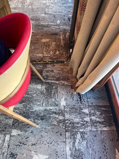 The image depicts an indoor scene, likely a room in a building in Val-d'Isère, France.
In the foreground and midground, a modern chair is visible, partially obscured. It features a bright red backrest and a cream-colored seat or cushion, with light wooden legs extending towards the bottom and right.
Towards the upper part of the frame, a window with a dark brown frame is barely visible at the very top. Below this, a light beige or tan pleated curtain or blind is partially drawn. Underneath the curtain mechanism, a dark structure, possibly a shelf or part of the blind's housing, is visible. This area also reveals black cables and a white connector, suggesting an electronic or powered system for the blind. A piece of fabric with a reddish and dark pattern can also be seen below the curtain.
The floor is covered with a distinctive patterned material, appearing as dark grey and black blocks with a distressed or abstract texture, resembling aged stone tiles or a contemporary carpet design.
No people are present, and no specific activity is taking place. The scene is static, showcasing interior decor elements. There is no visible text in the image. The overall impression is one of a modern and functional living space.