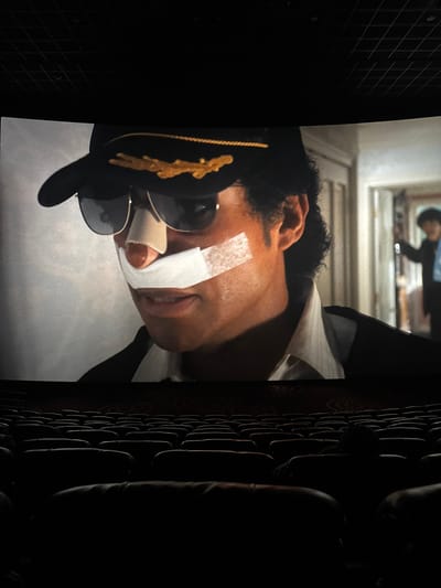 The image shows a close-up of a person lying on a white surface, possibly a bed or table. The person is wearing a black hat with gold detailing and sunglasses. There are bandages on their forehead and nose. In the background, there is a reflection in a window, and a textured wall on the right. The location of the scene is unknown; the image does not provide enough information to determine if it is in Taihe District, China, or anywhere else.