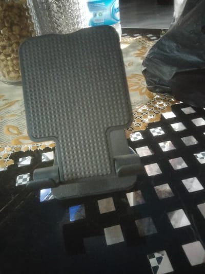 The image displays an indoor scene, likely set on a table covered with a patterned surface. The central object is a dark grey or black foldable stand, possibly for a phone or tablet, standing upright. Its surface features a fine, raised dot pattern for grip.

To the left of the stand, a clear glass jar filled with light brown, irregularly shaped snacks or nuts is visible. Behind the jar, a plastic water bottle, predominantly white and light blue, is partially obscured, with fragments of text like "KO" and "mineral" discernible on its label.

The table surface itself is layered. An underlying cream and orange/yellow patterned fabric or mat with floral motifs and intricate borders is visible. Overlapping or adjacent to this, particularly on the right and foreground, is a dark, reflective surface embedded with multiple square-shaped reflective elements. Some of these squares exhibit a black and white checkerboard pattern, while others show distorted reflections of the surrounding environment, including hints of blue.

On the far right, a crumpled black plastic bag rests on the table. No human subjects or explicit activities are present, portraying a still life arrangement of household items. The lighting appears consistent with an indoor setting, with no specific time of day indicated.