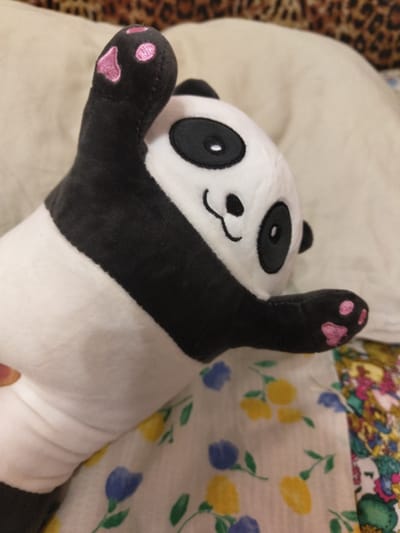 The image shows a close-up, eye-level shot of a plush panda toy lying on a light-colored surface, possibly a bed or sofa. The panda is positioned on its back with its legs extended. Its head is tilted slightly to the left, and its arms are raised. The panda has the typical black and white markings, with black eye patches, ears, and a band across its body. Its paws have pink embroidered paw prints.
In the background, to the left of the panda, is a textured surface with a leopard print pattern. To the right of the panda, a floral patterned fabric is visible, featuring small yellow and blue flowers with green stems on a white background.
The lighting suggests an indoor setting, possibly during the daytime given the brightness. There are no visible signs of a specific event or activity taking place, other than the panda being positioned as if it were relaxing or playing. The overall mood is playful and relaxed. No text is visible in the image. The location context is Melitopol, Ukraine.