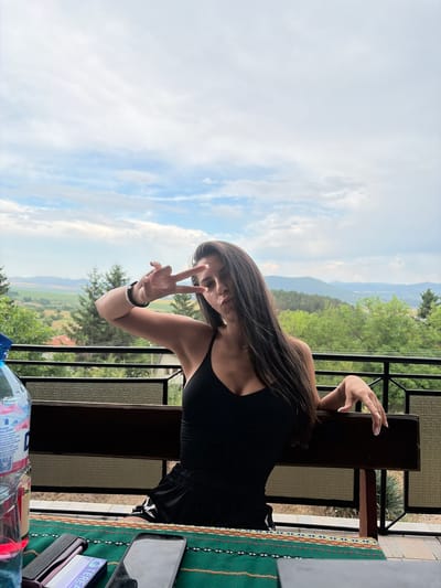 A young woman with long, dark hair is seated on a wooden bench on a balcony, outdoors. She is wearing a black tank top and black shorts. She is making a peace sign with her right hand, holding it near her face. A bottle of water and a smartphone are visible on a table in front of her, covered with a colorful textile. The background shows a scenic view of rolling hills, trees, and a cloudy sky, consistent with the landscape around Vladislavtsi, Bulgaria. The railing of the balcony is visible in the foreground and mid-ground.