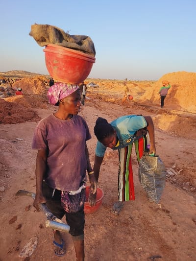 This image depicts an outdoor scene in the city of Shen, Nigeria, at what appears to be a manual excavation or mining site under a clear, pale blue sky. The ground is reddish-brown, dry, and dusty, with numerous large mounds of earth visible across the wide landscape.
In the foreground, two adult females are engaged in labor. The woman on the left, with dark skin, wears a dusty light purple t-shirt, dark trousers, blue flip-flops, and a patterned pink and purple headscarf. She balances a large, red plastic basin containing a brown burlap sack on her head. In her right hand, she holds a hoe-like tool with a metallic handle and a silver cylindrical object, possibly a can or bottle. Her gaze is directed towards the right side of the frame.
To her right, slightly behind and bent forward, is another adult female with dark skin. She wears a light blue short-sleeved top with a faint design, brightly colored vertically striped pants (red, green, yellow, black), and grey flip-flops. Her left hand holds a black plastic bag, partially displaying the word "SHOPPING," while her right hand is extended towards a red plastic bucket on the ground.
In the mid-ground and background, additional indistinct figures are seen scattered among the earth mounds, also appearing to be engaged in similar manual tasks. One figure on the far right carries a red basin on their head. A white vehicle is partially visible further back among the mounds.