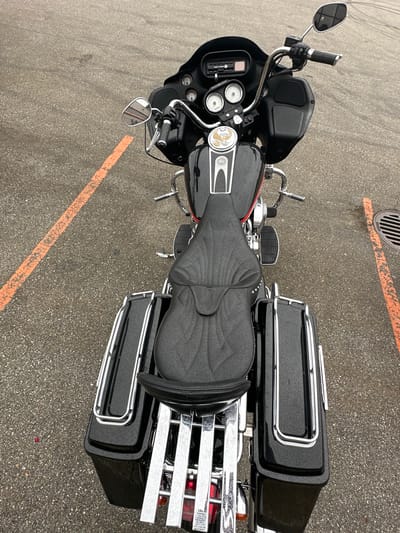 A top-down, slightly angled, full shot captures a black motorcycle parked on asphalt. The motorcycle is a touring model with a large front fairing, saddlebags, and a passenger backrest. It has a black quilted seat with decorative studs. Chrome accents are visible on the handlebars, mirrors, saddlebag lids, and a luggage rack at the rear. The motorcycle is oriented diagonally across the frame.
The ground is paved asphalt with visible orange painted lines, suggesting a parking lot or road marking. A portion of a metal grate, likely a drain cover, is visible in the upper left quadrant of the image. The lighting suggests it is daytime, and the speckled appearance of the asphalt indicates it may be wet, possibly from recent rain or dew.
No people are visible in the image. The scene is static, with the motorcycle stationary. The location context is the Township of Langley, Canada, though no specific landmarks are identifiable within the image itself beyond the generic parking lot setting. There is no visible text in the image.