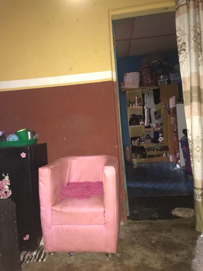 An interior view in Oba-Ile, Nigeria, depicts two adjoining rooms. The foreground room features a bi-color wall, with an upper yellow section and a lower reddish-brown section, separated by a white horizontal stripe. The concrete floor is soiled. A black compact refrigerator or cabinet stands on the left, adorned with pink floral stickers. On its top rests a green plastic tray containing various containers, including a blue plastic sieve. To the right of the refrigerator is a pink single armchair, over which a folded red or pink fabric is draped. The armchair is supported by four metallic legs. Beneath the refrigerator, a black and white patterned mat is visible.
An open doorway on the right leads to a second room. A striped curtain, beige and white, partially obscures the doorway on the right. The second room has blue walls and a darker floor, possibly tiled. A wooden shelving unit or wardrobe dominates the left side of this room, containing numerous items including bottles, bags, and shoes on shelves, and various garments hanging from a rail. Additional bags and containers are stacked on top of the shelving unit. The ceiling of this inner room reveals exposed wooden beams. No individuals are present in either room, and no actions or interactions are occurring.