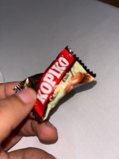 A hand holds a Kopiko candy wrapper against a light-colored fabric backdrop. The wrapper is red, yellow, and brown.