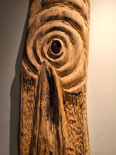 A vertical, close-up shot captures a carved wooden art piece mounted on a wall. The wood is light brown with darker striations and appears textured, with some areas exhibiting rough, almost splintered surfaces. The art piece itself is abstract and features carved concentric circles, reminiscent of a ripple effect or an eye, with a dark, hollow center. Below this central design, the carving descends into a more jagged, organic pattern. The piece is lit from the front, casting a shadow on the left side of the wall, which is a muted gray. The wall to the right of the art piece is a pale cream color. The scene is indoors, and based on the style and context of art often found in tourist areas, this artwork could be displayed in a gallery, hotel lobby, or a restaurant in the city of Labuan Bajo, Indonesia, known for its natural beauty and growing tourism infrastructure. There are no people or animals visible in the image.