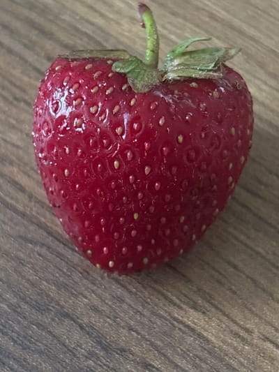 This is a close-up, overhead shot of a single, ripe strawberry placed on a wooden surface. The strawberry is a deep red color with numerous small, yellowish seeds embedded in its skin. The surface it rests on appears to be made of wood with a visible grain pattern running diagonally from the top left to the bottom right. The lighting suggests it is daytime, possibly indoors with natural light, as there are subtle highlights on the strawberry's surface indicating a gentle illumination. No people are visible in the frame. The context of Pereiaslav, Ukraine, does not provide any specific visual cues within the image itself, but it places the scene geographically. The image focuses solely on the fruit and its immediate surroundings, with no indication of any particular activity or event.