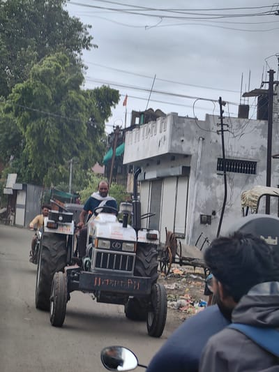 The image captures a street scene in Malkapur, India. In the foreground, a white tractor with large rear wheels is being driven by a man wearing a blue shirt and a white scarf around his neck. The tractor has a black front bumper with text in orange. Behind the tractor, another man is riding a motorcycle, wearing a yellow shirt. The street is paved, and on the right side of the image, there is a building with a gray, weathered facade and shuttered windows. Adjacent to the building, a utility pole with numerous wires is visible. The environment appears to be a moderately populated urban or semi-urban area, with some greenery from trees in the background. The sky is overcast. There is debris and trash scattered on the ground near the right side of the street. The image is taken from a slightly elevated perspective, possibly from another vehicle on the road.