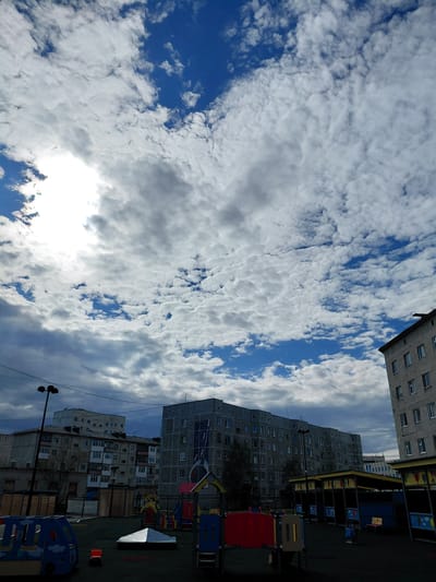 The image captures an urban residential scene in Nadym, Russia, under a dynamic sky. The sky dominates the upper and central portions of the photograph, featuring bright blue patches interspersed with a wide array of white and grey clouds, suggestive of a partly cloudy day. A bright area on the left hints at the sun's location, partially obscured by clouds.
In the lower and right parts of the frame, several multi-story apartment buildings are visible. These structures appear to be concrete or stone, with numerous windows. One prominent dark grey building in the mid-right features a large mural or artistic design on its side, facing the viewer. Adjacent to and in front of these buildings is a children's playground, equipped with colorful structures in red, blue, and yellow, likely including slides or climbing frames. The ground of the playground appears to be a dark, possibly artificial surface.
Additional objects include a black streetlight pole with multiple lamps positioned on the left side, and several overhead utility wires stretching across the sky, connecting to the buildings. The overall setting depicts a typical daytime scene in a residential district, characterized by Soviet-era architectural influences common in Russian cities. No people or discernible text are visible in the image.
