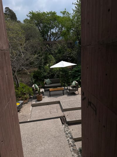 The image depicts a scene in Tepoztlán, Mexico, from an elevated, interior perspective looking outwards. The view is framed by a rough concrete structure, likely part of a building. Outside, a patio is visible, furnished with modern outdoor seating including a sofa, chairs with patterned cushions, and a wooden coffee table. A white sun umbrella provides shade. The patio is surrounded by lush greenery, including various shrubs, trees, and a glimpse of a larger forest area. The patio surface consists of large, irregularly shaped concrete slabs with gravel in the gaps. The sky is overcast.
