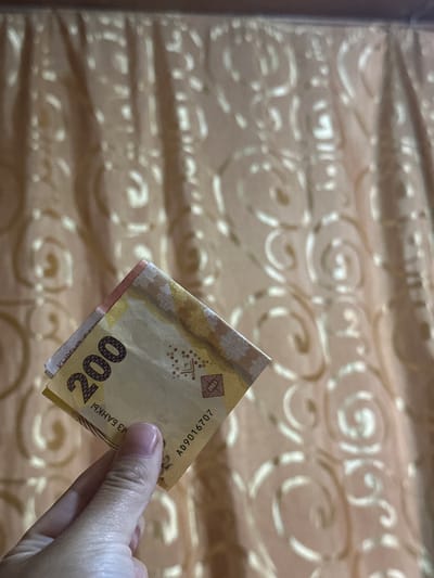 A close-up, high-angle shot shows a hand holding a folded banknote in front of a beige, patterned curtain. The banknote is predominantly yellow with red and blue accents, and prominently displays the number "200". Text on the banknote is partially obscured but includes "2023" and "AD9016707". The background curtain has a repeating swirl pattern. The scene does not provide any information regarding a specific city or location.