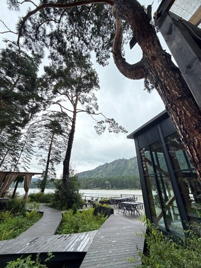 This image depicts an outdoor seating area adjacent to a modern building, situated in a natural environment with a river and forested hills in the background. A winding wooden walkway leads through lush greenery towards the seating area. The seating area features tables and chairs, and string lights are visible overhead. The building itself has large glass windows and a dark facade. The sky is overcast. The location is identified as Nizhnekayancha, Russia.