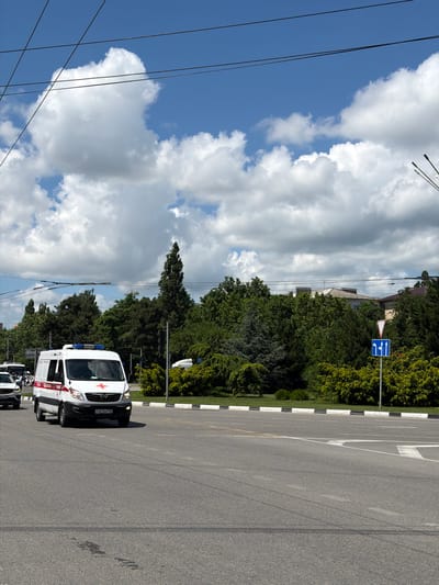 An ambulance with flashing blue lights and a red cross emblem is driving on a paved road in Abinsk, Russia. The ambulance has the license plate "046KK 193". Several power lines are visible overhead, and lush green trees line the side of the road. A traffic sign with arrows indicating lane direction is also visible. The sky is blue with fluffy white clouds.