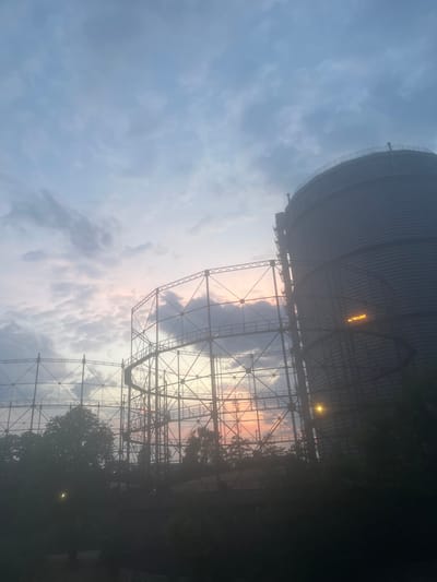The image captures a sunset view of the Augsburg gasworks in Augsburg, Germany. The sky is filled with scattered clouds tinged with the soft hues of pink and orange from the setting sun. Silhouetted against the sky are two prominent structures: a large, cylindrical gas holder on the right and a latticed framework of what appears to be another gas holder on the left. The latticed structure is intricate, with visible scaffolding and walkways. Trees and vegetation are visible in the foreground and midground, silhouetted against the lighter sky. A bright, yellowish light source is visible on the cylindrical gas holder, and another is present near the bottom left of the image. The overall atmosphere is muted and slightly hazy, typical of twilight conditions. There are no discernible people or animals in the frame.