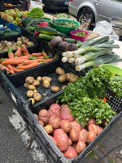 The image shows a street market stall displaying a variety of fresh produce in black plastic crates. The stall is set up on a paved surface, possibly a sidewalk or street, in Senhora da Hora, Portugal, as indicated by a license plate on a car in the background.
Dominating the foreground are several crates filled with different vegetables. One large crate is overflowing with vibrant orange carrots, their green tops still attached. Adjacent to the carrots are crates of potatoes, some lighter brown and smaller, others larger and reddish-pink. A generous bunch of fresh, leafy green parsley is also prominent, spilling out of a crate. Other vegetables visible include beets, leeks, green beans, cucumbers, peppers, and onions.
In the background, a silver car is parked, and some of the produce is arranged on a table or stand. The overall impression is one of a bountiful and fresh selection of vegetables offered at a local market. There are no people visible in the image.