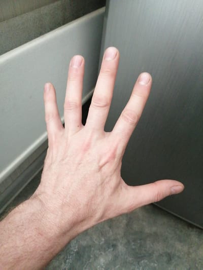 A person's left hand is extended towards the camera, palm facing upwards. The hand is positioned against a blurred background consisting of a light gray surface with vertical lines and a darker gray surface. The fingers are spread open, with the thumb slightly separated from the other fingers. The fingernails appear to be short and unpolished. The skin on the back of the hand shows visible veins and some light hair. There is no visible indication of location within Norilsk, Russia, or any other contextual information that would allow for such a determination.