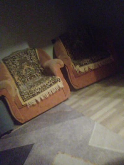 Two armchairs with patterned throws are visible in a dimly lit room. The armchairs appear to be upholstered in a reddish-orange fabric and are decorated with intricately patterned throws that have fringed edges. One armchair is positioned in the foreground on the left, and another is in the mid-ground on the right. The flooring is light-colored wood with visible plank lines, and a light gray rug or carpet is partially visible in the lower left corner of the image. The background is dark and indistinct, suggesting poor lighting conditions or shadows. There are no people or overt actions depicted in the scene. The location is identified as Shakhtinsk, Kazakhstan.