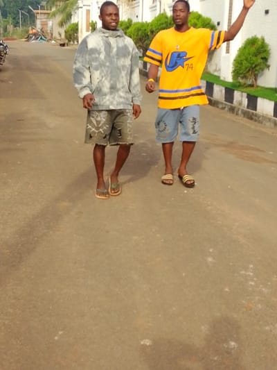 Two men stand on a paved street. One wears a grey and white hooded sweatshirt and patterned shorts. The other wears a yellow and blue t-shirt with jean shorts. Both have on sandals. The man in yellow raises his arm. A building sits behind them on the right, and a street with buildings is behind them on the left. Green bushes are to the right of the men.