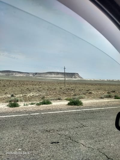 This media file captures a view from inside a vehicle looking out at a vast, arid landscape under a cloudy sky. The foreground shows a cracked asphalt road with a white line marking its edge. To the left of the road, sparse, dry vegetation and some green bushes are visible. In the middle ground, a wide, flat expanse of dry grass and scrub stretches towards a distant mesa. A single utility pole with wires stands in the middle distance, adding a man-made element to the natural scenery. The background is dominated by a large, layered mesa with a distinctive, eroded shape, displaying shades of gray, white, and brown. The sky is overcast, suggesting it is daytime, likely morning given the timestamp. The overall mood is one of travel through a remote, natural environment.
The visible text reads "2026/06/26 10:47", indicating the date and time the image was captured. The location context provided is Saiyn Şapağatov, Kazakhstan, which aligns with the arid, steppe-like terrain depicted. There are no people or specific objects other than the road and utility pole that would suggest a particular activity or event beyond traveling through the landscape.