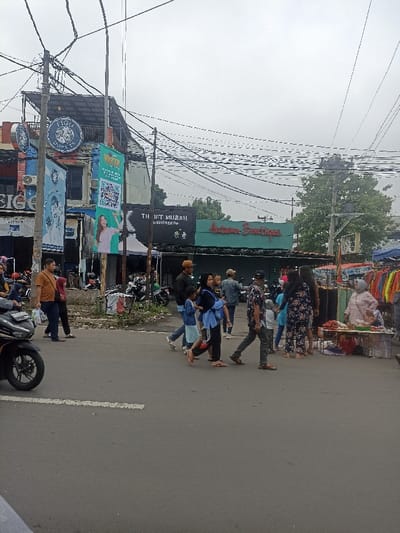 The scene is a street in Purwokerto, Indonesia, located at latitude -7.417 and longitude 109.249. The street is lined with buildings on both sides, including a shop called "Thrift Murah" and a shop called "Astana Boutique." There are several people walking along the street, some carrying bags or packages. There are also several motorcycles parked on the side of the road. In the background, there is a busy street intersection. The sky is overcast and gray, suggesting that it may be raining or about to rain. The scene is a typical street scene in a busy Indonesian city.
