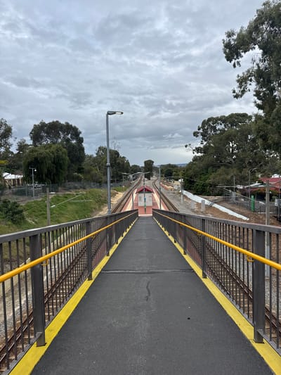 The image depicts a railway overpass in Bassendean, Australia. A straight, asphalt pathway with yellow safety edges runs between metal railings, leading to a train station platform in the distance. Parallel train tracks extend from the platform into the distance. Adjacent to the tracks are grassy areas with trees and utility poles. The sky is overcast with cloudy skies. There are no people visible in the scene.