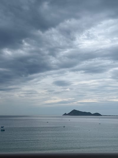 The image depicts a serene seascape at the city of Song Cau, Vietnam. The sky is overcast with dramatic, dark clouds that occupy the upper half of the frame. Below the clouds, the sea stretches out, a vast expanse of calm, gray-blue water. Several small boats are scattered across the water, some appearing as dark silhouettes against the lighter surface. In the distance, a prominent, dark island with a rugged silhouette rises from the sea, its form softened by the hazy atmosphere. The horizon line is clearly defined, separating the sea and sky. The foreground is occupied by a dark, out-of-focus railing, suggesting the vantage point is from a balcony or elevated structure. The overall mood of the image is tranquil, yet tinged with the somber beauty of a cloudy day.