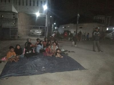 The image captures a nighttime scene in Baleendah, Indonesia, showing a group of people gathered outdoors on what appears to be a street or open paved area. The primary subjects are a diverse group of individuals, predominantly children and women, sitting close together on a large, dark blue tarp laid on the ground in the foreground. They are mostly facing towards the right side of the frame, some appearing to look off-camera, while others are interacting amongst themselves. Their expressions are generally subdued or attentive, though difficult to discern clearly in the low light.

To the right of this seated group, a man wearing a cap and a jacket stands, engrossed in his mobile phone. Further in the background, a small dog and several other individuals, including adults and children, are standing along the side of the road. The setting is illuminated by various artificial lights, including streetlights and lights from nearby houses, creating bright spots and deep shadows.

In the background to the left, a house with visible windows and a metal gate can be seen, behind which a silver car is parked. Other residential buildings are visible in the distance, further suggesting a neighborhood setting. The overall activity suggests a communal gathering, possibly for watching an event, seeking temporary refuge, or simply socializing in the evening. The time of day is clearly night, characterized by the darkness and reliance on artificial illumination. No visible text is clearly legible within the image.