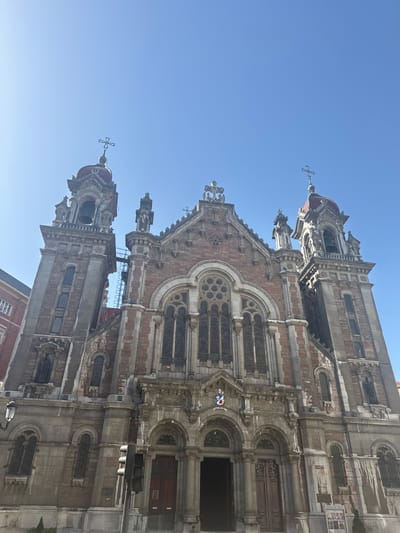 This media file displays the facade of a grand church, its intricate architecture reaching towards a clear blue sky. The building is constructed with a combination of light-colored stone and reddish-brown brick, featuring multiple towers, ornate carvings, and arched windows. Several crosses are visible atop the church's spires, indicating its religious purpose.

The image appears to be taken during the day, under fair weather conditions, given the bright sky and strong sunlight casting shadows on the building. The perspective suggests the viewer is looking up at the church, emphasizing its height and grandeur.

One section of the building has a small, colorful mural of what appears to be a child dressed in blue and red, waving with a white glove, next to a flag, and the letters "SANT" are partially visible below it. This detail adds a touch of modern street art or decoration to the historic structure.

The architecture itself suggests a Gothic or Neo-Gothic style, common in European religious buildings. The detailed stonework and the presence of bells in one of the towers are notable features. While no specific activity is clearly depicted, the scene captures the timeless presence of the church within what is likely an urban setting. The clear sky and sunlight suggest a pleasant day. The location context provided, Oviedo/Uviéu, Spain, helps to place the building geographically.