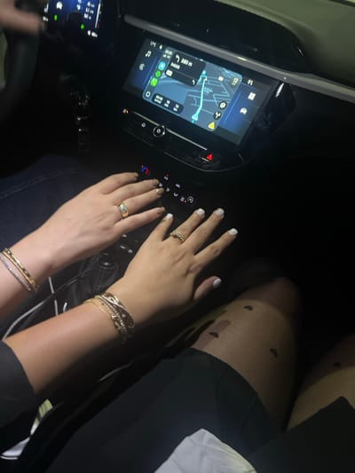 Inside a car at night, two pairs of hands rest near the dashboard, one with white nail polish and a beaded ring, the other with gold nail polish and a wide gold ring. The first hand displays multiple gold bracelets. A navigation screen glows on the dashboard. A person wears tights with small heart patterns.