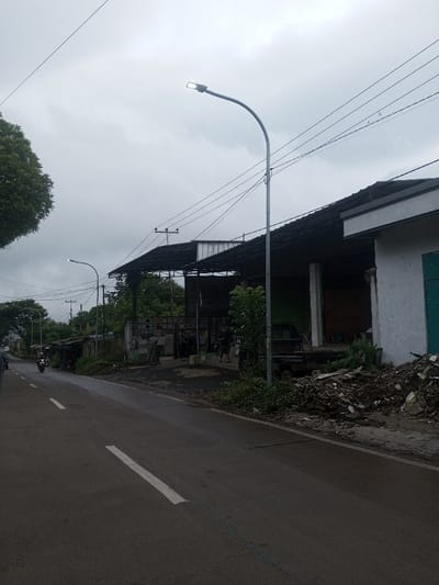 A street scene showcases a wet, dark asphalt road with a white lane marker. Buildings with dark roofs line the right side of the road, accompanied by a streetlight. A motorcycle drives on the left side. Debris and vegetation clutter the roadside. Overhanging electrical lines stretch across an overcast sky. People stand near the buildings.