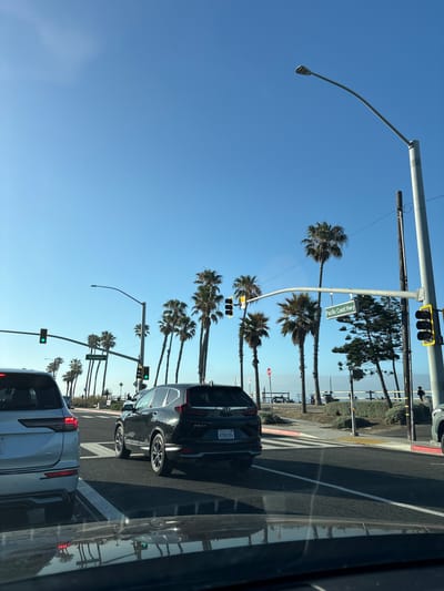 The image depicts a street scene in Huntington Beach, California, USA, characterized by palm trees lining the thoroughfare under a clear blue sky. A street sign indicates "Pacific Coast Highway." Several vehicles are visible, including a silver SUV and a dark-colored sedan, stopped at a traffic light that is currently green. The road markings denote a crosswalk. The image appears to have been taken from inside a vehicle, likely another car or truck, due to the presence of the vehicle's window frame on the right side of the shot.