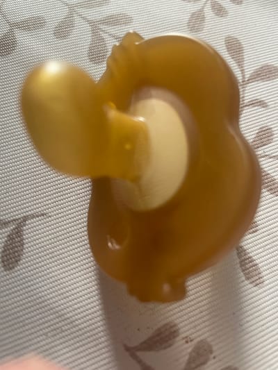 A close-up, slightly out-of-focus, overhead shot displays a single, amber-colored pacifier on a textured, white and brown patterned surface. The pacifier is positioned on its side, with the nipple extending to the left. The surface appears to be a placemat or tablecloth with a floral design. The image does not provide any information or context related to the city of Otradnoye, Russia, or any other specific geographical location.