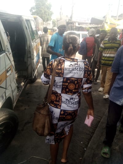 A woman wearing a dress patterned with "Hermes Paris" logo stands in a busy street scene. Several people surround her, some standing near a white van with an open door. A man in a blue shirt is visible in the background. The woman carries a brown bag over her shoulder and holds a pink piece of paper in her hand. The image depicts a typical urban setting with people and vehicles amidst buildings and advertisements.