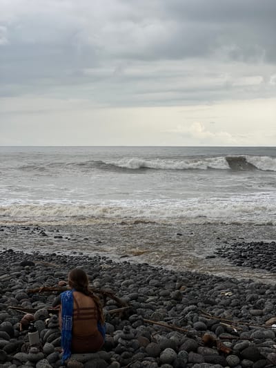A lone individual sits on a rocky beach in El Tunco, El Salvador, facing the ocean. The sky is overcast with a predominantly grey and white color palette. The sea is choppy with white-capped waves breaking near the shore. The beach is covered in smooth, dark grey and black stones of varying sizes. The individual is wearing a brown bathing suit and has a blue scarf draped over their shoulders. Their hair is braided and falls down their back. A white water bottle and some pieces of driftwood are visible on the beach near the person. The scene conveys a sense of solitude and observation of the natural environment.