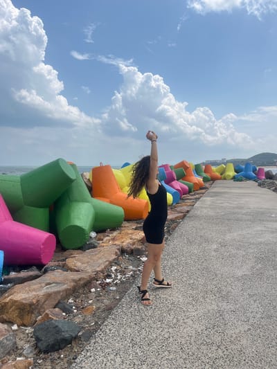 In the city of Phú Thủy, Vietnam, a woman with curly dark hair stands on a paved walkway next to a breakwater made of colorful tetrapods. The woman is wearing a black tank dress and sandals. Her arms are raised above her head, and she appears to be stretching or posing. The tetrapods, a type of concrete armor unit, are painted in bright colors including pink, green, orange, yellow, and blue. They are arranged along the coastline, protecting it from the sea. The sea is visible in the background, with a hazy shoreline and buildings in the distance. The sky is partly cloudy with large, white cumulus clouds against a blue sky. The ground on the left side of the walkway is rocky and covered with debris.
