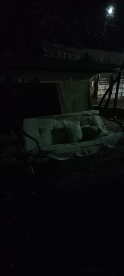 In the city of Iskra, Bulgaria, a dark scene unfolds, illuminated by a solitary light source. A green swing set, draped with a matching canopy, occupies the foreground. The swing's cushioned seat, a lighter shade of green, features two tufted pillows. Behind the swing, a dark green fabric hangs, partially obscuring a metal fence. The ground around the swing is covered in dark soil. The overall atmosphere is one of stillness and shadow.