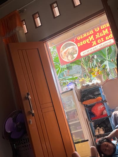 The image depicts a slightly tilted indoor scene, likely a domestic setting or a small shop in Serdang Kulon, Indonesia. A wooden door with a silver handle dominates the left side of the frame. Sunlight streams in from an open doorway, illuminating a red banner with yellow text and a picture of food. The banner appears to be advertising a food establishment, with text that is partially obscured and reversed. Beyond the doorway, lush green foliage suggests an outdoor or greenhouse environment.
Inside the room, a purple fan is visible on the left. To the right, a white mini-fridge stands next to a blue shelving unit filled with various items, including plastic bags, shoes, and what appear to be food containers. In the foreground, a person is lying down, with their head resting on their hand and their hair spread out. Their face is partially visible. The overall impression is one of a casual, everyday scene, possibly a home or a small business, bathed in the warm light of the afternoon sun. The presence of a fan, a fridge, and a food banner suggests a place where people live and perhaps sell food.