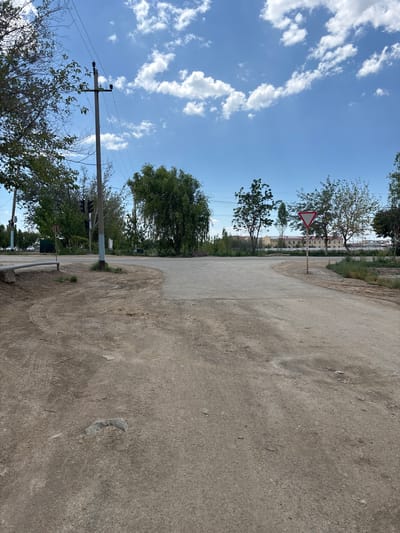 The image depicts a street intersection in Nukus, Uzbekistan. A paved road curves to the right, with a dirt and gravel shoulder on the left. A traffic light and utility pole are visible on the left side of the road. A yield sign is present in the middle of the intersection. Trees line the sides of the road, and buildings can be seen in the background. The sky is blue with scattered clouds. Tire tracks are visible in the dirt on the left side of the road.