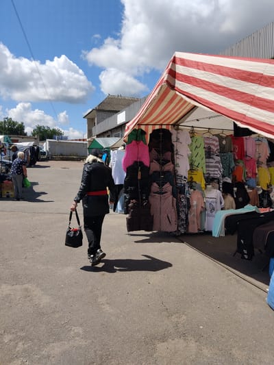 A blonde woman in a black jacket, black pants, and sneakers walks away from the camera carrying a black bucket bag. She has a red belt around her waist. She is walking on an asphalt path at an outdoor market with stalls set up on the right side of the image. The stalls are covered by striped red and white awnings and display various clothing items, including t-shirts, jackets, and pants. In the background, other market stalls and buildings are visible under a bright blue sky with scattered white clouds. The scene appears to be in the city of Chudniv, Ukraine, given the context provided.