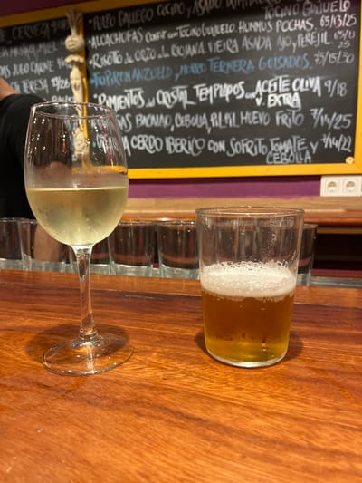 The image shows a wooden table with a glass of white wine and a glass of beer on it. The table is in a restaurant in Donostia-San Sebastian, Spain, located at latitude 43.324 and longitude -1.985. There is a black chalkboard menu with white chalk writing above the table with a purple wall behind it. Behind the table are several empty glasses. There is an arm and a hand partially visible in the image.  The glasses of wine and beer are the only drinks present on the table.  The restaurant's interior is visible in the background.