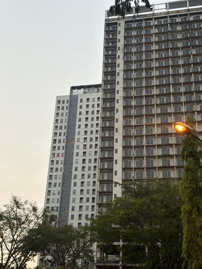 This image depicts two tall, Brutalist-style apartment buildings in Entalsewu, Indonesia. The buildings are characterized by repetitive, grid-like patterns of windows and concrete balconies. The first building, on the left and extending upwards, is darker and shows signs of age, with a large number of windows visible. The second building, to the right and lower down, is lighter in color and features a decorative band of clover-like symbols. A street lamp with an illuminated orange light is visible between the two buildings, casting a warm glow. Lush green trees partially obscure the buildings, with one tree in the foreground showing dense foliage and another in the background with more sparse branches. The sky is overcast, suggesting a cloudy or possibly hazy day. The image is taken from a low angle, emphasizing the scale of the buildings. No people are visible. The scene appears to be an urban residential setting.